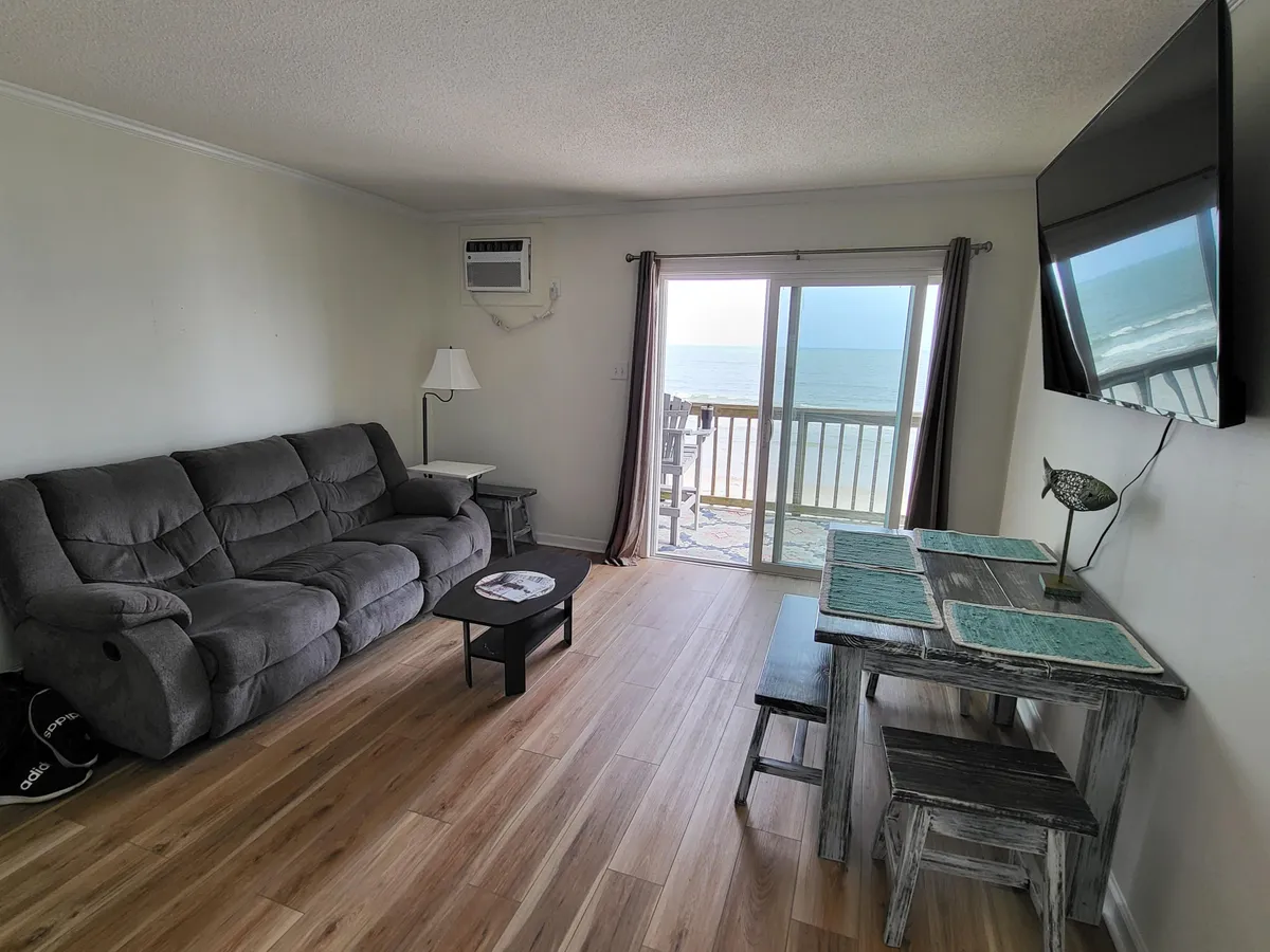 A cozy interior living and dining area featuring a sofa, dining table, and a stunning ocean view through a large sliding glass door. The scene is bright and serene, emphasizing the direct connection between the indoor space and the shoreline just outside.