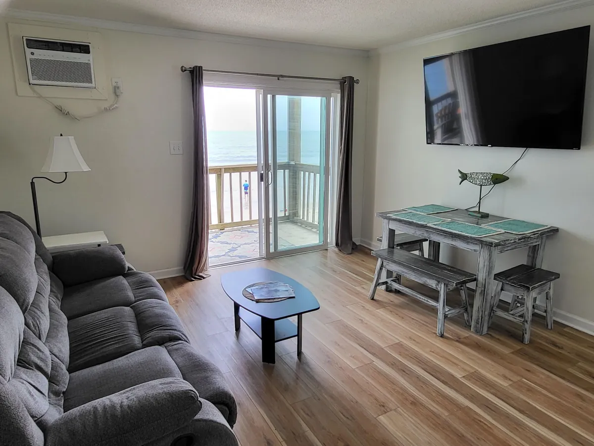 A serene living room with comfortable plush seating and a dining table, bathed in natural light streaming through large sliding glass doors. The doors open to reveal a breathtaking ocean view, blurring the line between indoor comfort and outdoor beauty.