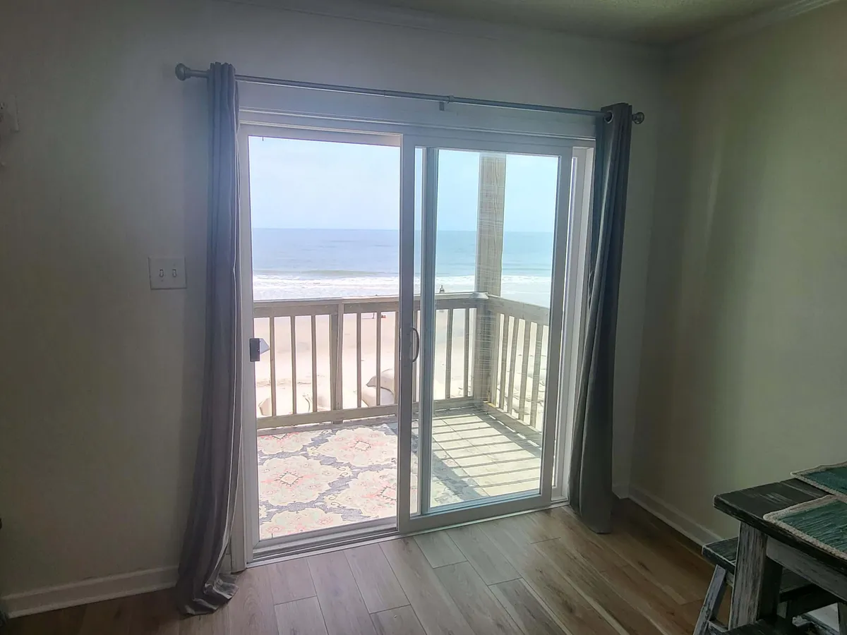 An inviting interior scene centered on expansive sliding glass doors overlooking the ocean. Beyond the doors, a wooden deck invites guests to relax and socialize while embracing sunrise views and the tranquil sounds of the waves.