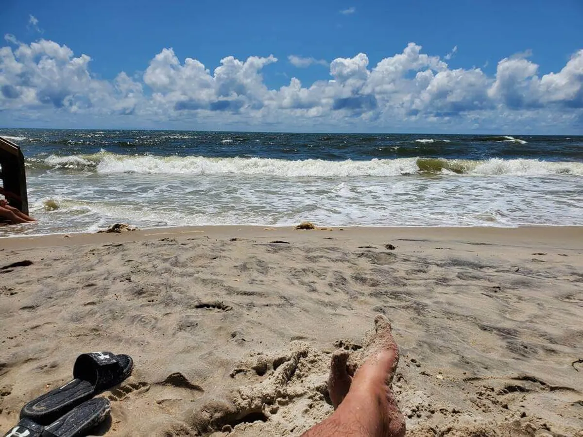 A tranquil snapshot of sandy beach with gentle ocean waves, a cloudy sky, and a pair of feet resting at the edge of the frame. The image exudes peace and solitude, drawing attention to the experience of relaxing at the shore and enjoying the surf.