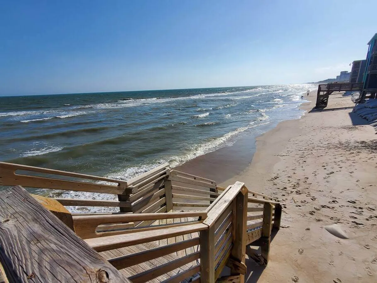 A set of wooden stairs leads directly from the property onto a sandy beach, with ocean waves gently rolling in the background. The inviting path underscores the proximity of the home to the shoreline, with easy, unobstructed access.
