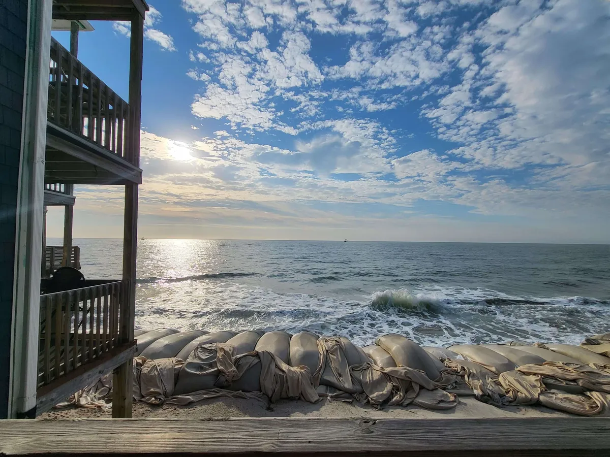 This serene view highlights ocean waves rolling onto the shore, sunlight reflecting on the water, and a wooden structure suggesting direct access to the beachfront. The cloudy sky completes the mood, hinting at peaceful mornings or evenings spent together by the sea.