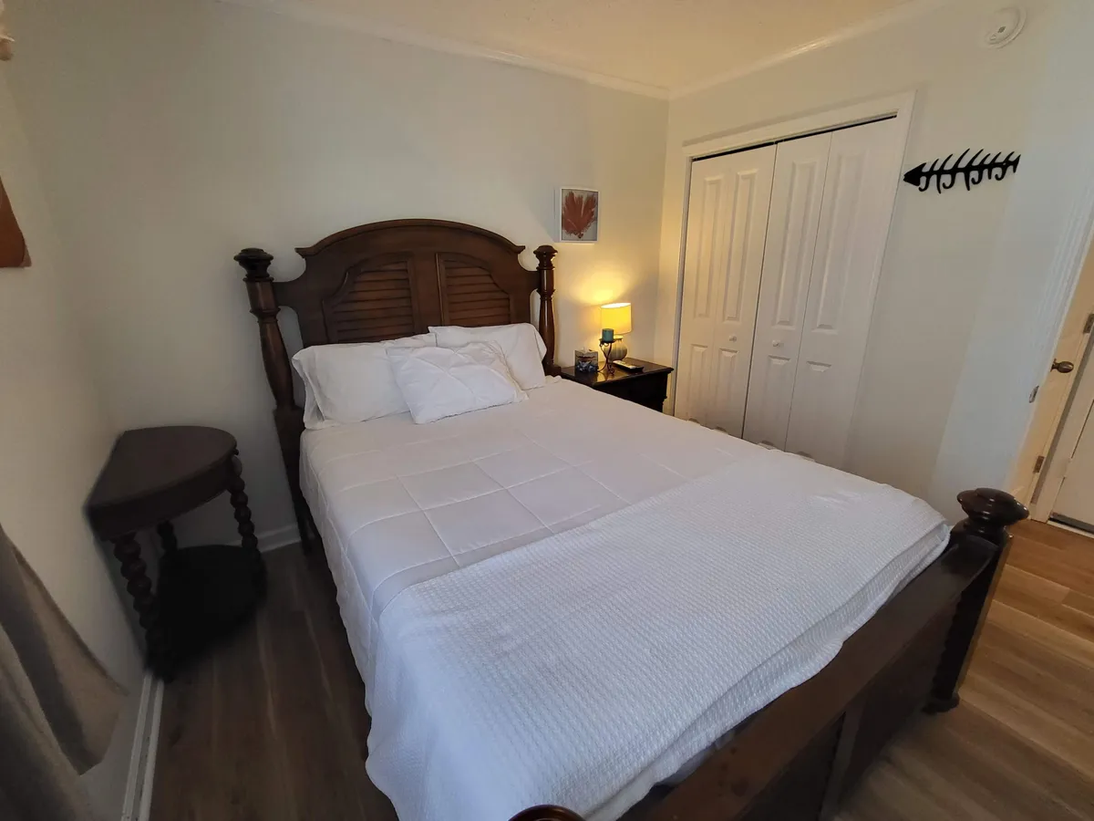 A peaceful bedroom with a wooden bed frame, crisp white bedding, a nightstand with a lamp, and closet doors. The scene is bathed in gentle natural light, creating a sense of calm and comfort ideal for unwinding or beginning your day.