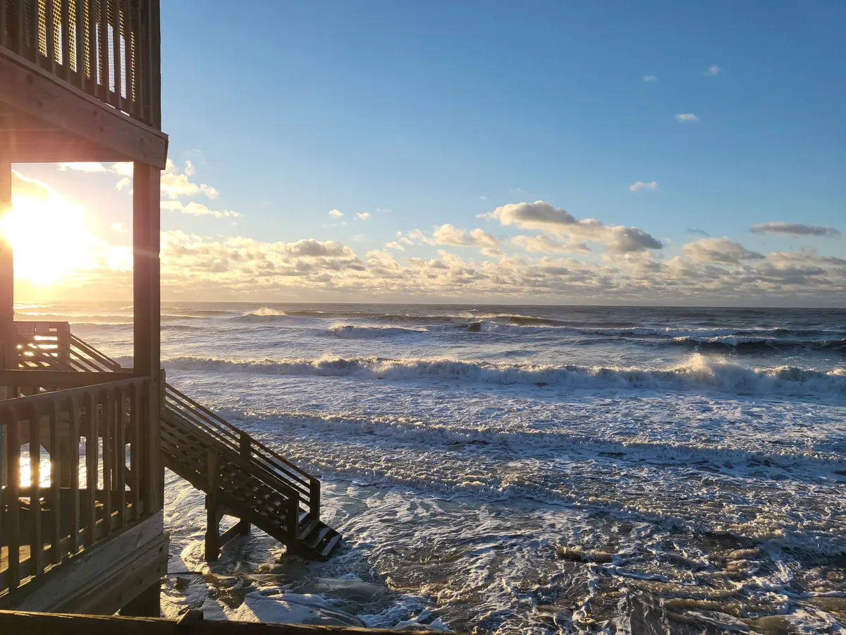 A wide view of the ocean at sunset, with waves crashing on the shore and a wooden stairway leading down to the beach. The atmosphere is serene and inviting, capturing the magic of beachside living and the convenience of immediate access.