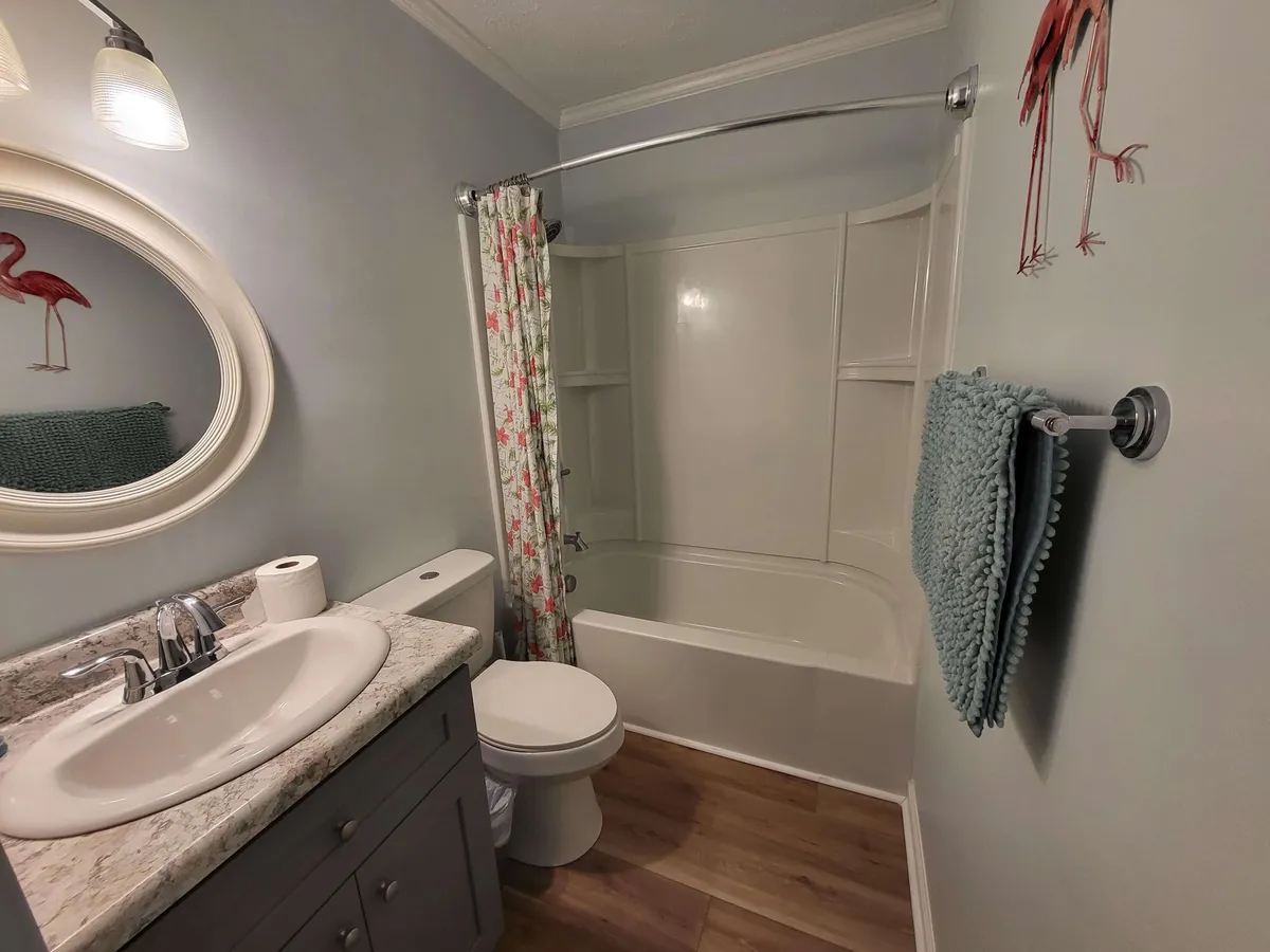 A newly remodeled bathroom scene with a modern sink and round mirror, a bathtub adorned with a floral shower curtain, and tasteful decorative elements including a towel and flamingo art. The space is bright, clean, and welcoming.